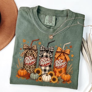 May include: An olive green t-shirt featuring a graphic of three Dr Pepper cans with straws and bows. The cans have leopard print, black and white buffalo plaid, and orange leaf patterns. Pumpkins and sunflowers surround the base of the cans.