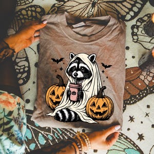 A person is holding a t-shirt with a Halloween-themed graphic design featuring a raccoon wearing a ghost costume, holding a cup with a pumpkin and a bat.