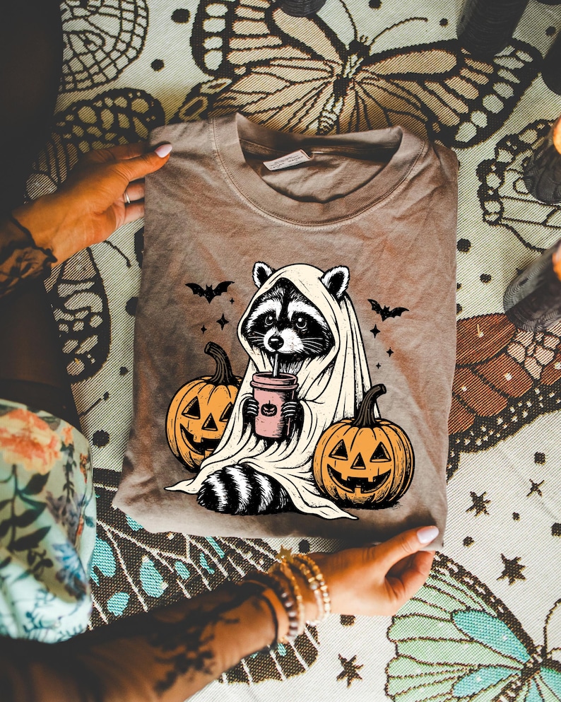A person is holding a t-shirt with a Halloween-themed graphic design featuring a raccoon wearing a ghost costume, holding a cup with a pumpkin and a bat.