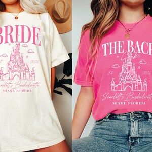 two women wearing t-shirts with the same design, which appears to be a wedding-themed graphic. The t-shirts are pink and white in color, and they both have the text "BRIDE" and "THE BACH" printed on them. The women are posing in front of a white wall, and they are wearing jeans and a hat.