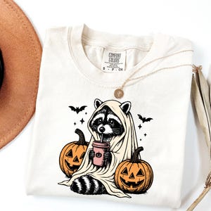 A white t-shirt with a graphic design depicting a raccoon dressed as a ghost, holding a cup, surrounded by jack-o-lanterns.