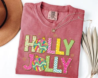 May include: A maroon Comfort Colors t-shirt with the words "HOLLY JOLLY" in colorful, patterned letters and Christmas ornament graphics. A silver necklace with a pendant is draped across the shirt. A brown hat is visible in the upper left corner.