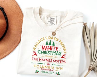 May include: Cream-colored t-shirt with a festive "White Christmas" design in red, green, and gold. The graphic includes text, Christmas trees, and reindeer. A gold necklace and a hat are visible.