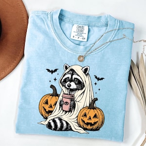 A blue t-shirt with a Halloween-themed graphic of a raccoon wearing a ghost costume and holding a pumpkin, accompanied by bats and a cup of coffee.