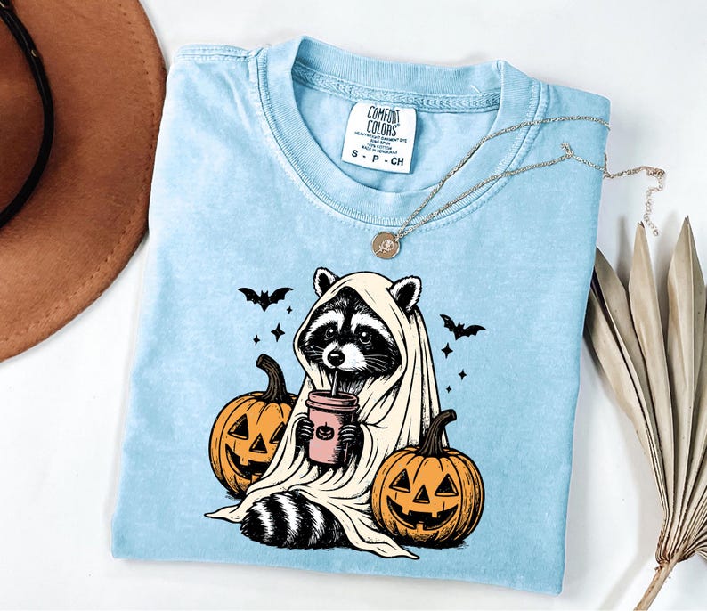 A blue t-shirt with a Halloween-themed graphic of a raccoon wearing a ghost costume and holding a pumpkin, accompanied by bats and a cup of coffee.