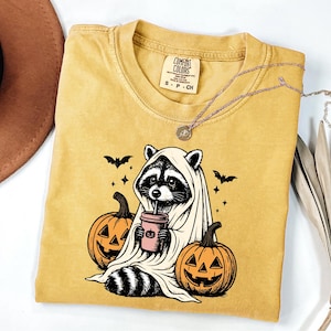 a yellow t-shirt with a Halloween-themed graphic of a raccoon dressed as a ghost, holding a cup of coffee and surrounded by carved pumpkins.