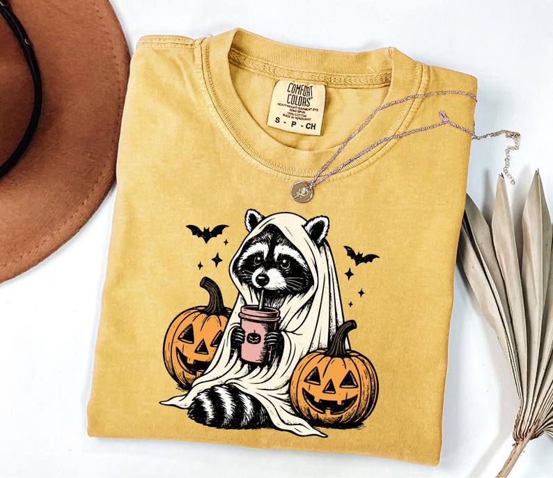 a yellow t-shirt with a Halloween-themed graphic of a raccoon dressed as a ghost, holding a cup of coffee and surrounded by carved pumpkins.
