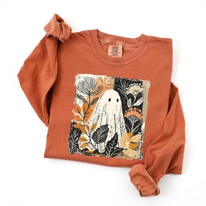 a long-sleeved orange shirt with a graphic design on the front, depicting a ghost surrounded by flowers and leaves.
