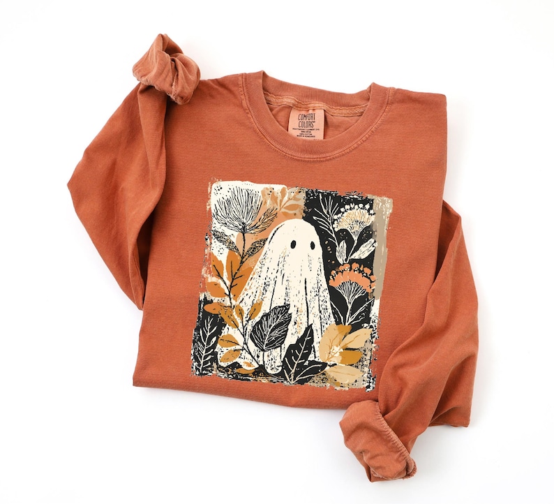 a long-sleeved orange shirt with a graphic design on the front, depicting a ghost surrounded by flowers and leaves.