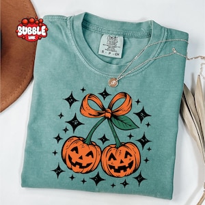 May include: A sage green crewneck t-shirt featuring a graphic of two orange jack-o'-lanterns connected by a stem with a large orange bow on top. The design is surrounded by black four-pointed stars. The shirt has a Comfort Colors brand neck label.