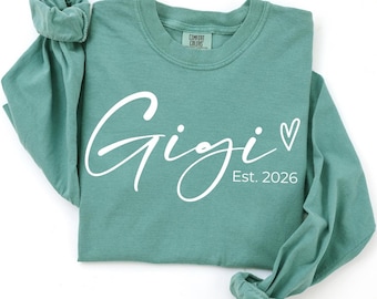 May include: A teal long-sleeve shirt with the word "Gigi" in white script, a small heart, and the text "Est. 2026". The shirt has a crew neck and rolled-up sleeves. The Comfort Colors brand tag is visible.