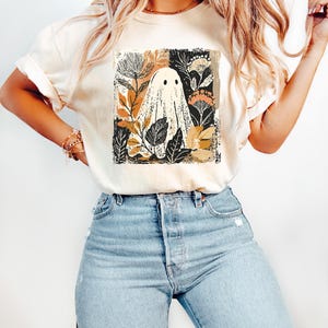 A woman wearing a white t-shirt with a graphic of a ghost surrounded by flowers and leaves, paired with blue jeans.