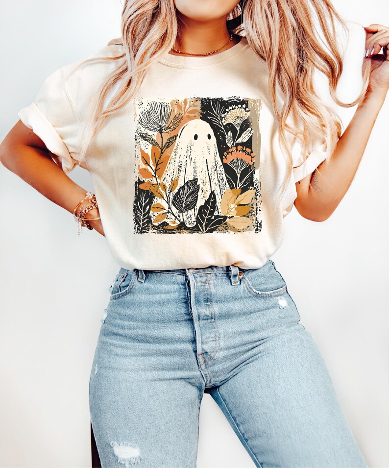 A woman wearing a white t-shirt with a graphic of a ghost surrounded by flowers and leaves, paired with blue jeans.