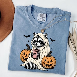 A blue t-shirt with a Halloween-themed graphic design, featuring a raccoon dressed as a ghost holding a coffee cup and surrounded by carved pumpkins.