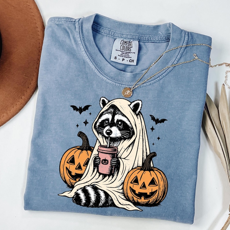 A blue t-shirt with a Halloween-themed graphic design, featuring a raccoon dressed as a ghost holding a coffee cup and surrounded by carved pumpkins.