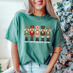 May include: A green short-sleeve t-shirt featuring a graphic of five distinct nutcracker figures in a row. Below the figures, the text reads THE NUTCRACKER CREW. The nutcrackers are decorated in various colors including green, red, pink, and black.