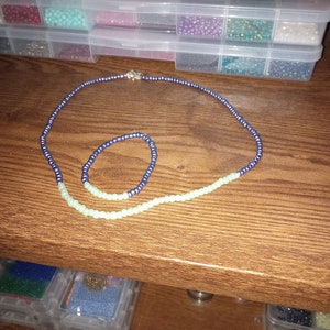 May include: Two beaded necklaces, one blue and one green, are displayed on a wooden surface. The necklaces are made of small, round beads. The blue necklace is longer than the green necklace.