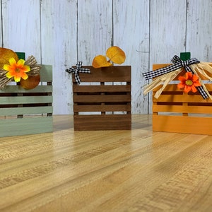May include: Three wooden pallet pumpkins decorated for fall. The pumpkins are painted green, brown, and orange. Each pumpkin has a different fall-themed decoration on top.