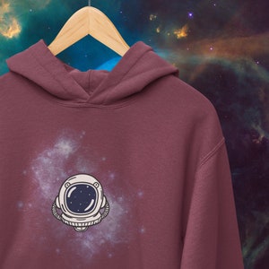 May include: A maroon hoodie with a white astronaut helmet graphic on the front. The graphic is surrounded by a white galaxy design.