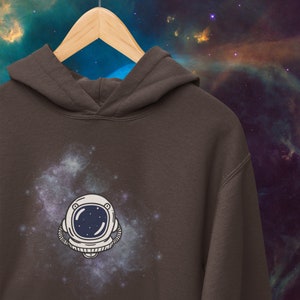 May include: A brown hoodie with a white astronaut helmet graphic against a galaxy background. The astronaut helmet has a dark blue visor and a white outline.