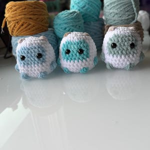 May include: Three handmade crochet creatures in white, blue, and green, with black eyes. Behind them are several skeins of yarn in shades of blue, green, and gold. The creatures have small, rounded ears and are sitting on a white surface.