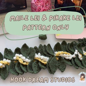 Op de afbeelding: Gehaakt Maile Lei & Pikake Lei patroon. De lei heeft donkergroene bladeren met witte en gele accenten. De tekst "MAILE LEI & PIKAKE LEI PATTERN ONLY" wordt weergegeven op een lichtgroene achtergrond. De tekst "HOOK DREAM STUDIOS" is ook zichtbaar.