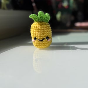 May include: A crocheted pineapple with a smiling face. The pineapple is yellow with a green leafy top and pink cheeks. The handmade fruit is a cute, whimsical design.