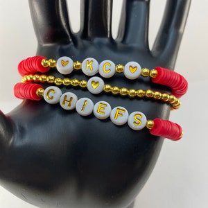 Könnte beinhalten: Drei gestapelte Armbänder mit roten und goldenen Perlen. Das obere Armband buchstabiert "K C" mit goldenen Buchstaben auf weißen Perlen und herzförmigen Akzenten. Das untere Armband buchstabiert "CHIEFS". Die Armbänder werden auf einer schwarzen Hand ausgestellt.