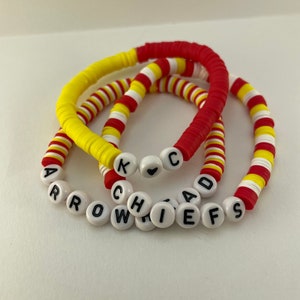 Könnte beinhalten: Drei Armbänder aus roten, gelben und weißen Perlen. Ein Armband buchstabiert "Chiefs" und "Arrowhead" mit weißen Buchstabenperlen. Die anderen Armbänder haben rote und gelbe Perlen. Die Armbänder sind gestapelt.