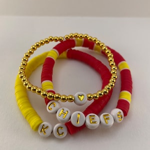 Könnte beinhalten: Drei Perlenarmbänder in Rot, Gelb und Gold. Ein Armband buchstabiert "CHIEFS" mit weißen Buchstabenperlen. Die anderen Armbänder sind einfarbig. Eines hat eine goldene Herzperle. Die Armbänder sind auf einer weißen Oberfläche gestapelt.