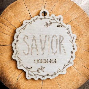 May include: Wooden ornament with a floral wreath design. The ornament says "SAVIOR" and "1 JOHN 4:14".