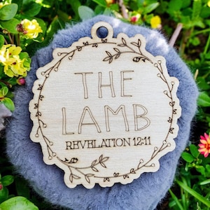 May include: A round wooden ornament with a floral design and the words "THE LAMB REVELATION 12:11" engraved on it.