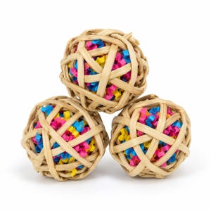 May include: Three spherical toys constructed from woven light tan natural reeds. Each ball features an open lattice design revealing a dense interior filling of small, crinkled paper strips in vibrant shades of bright pink, yellow, and blue.