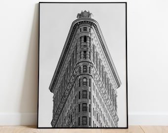 May include: Black and white framed print of the Flatiron Building in New York City. The iconic triangular building is centered in the frame, showcasing its architectural details. The print is displayed on a wooden floor against a white wall.