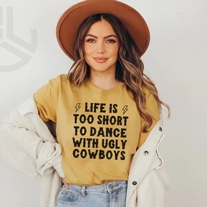 May include: A woman wearing a mustard yellow t-shirt with the text "Life is too short to dance with ugly cowboys" and a brown hat. She is wearing a white jacket and blue jeans.