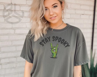 May include: A dark gray t-shirt with the text "Stay Spooky" in black lettering. A green zombie hand with red fingernails is making a rock and roll hand gesture.