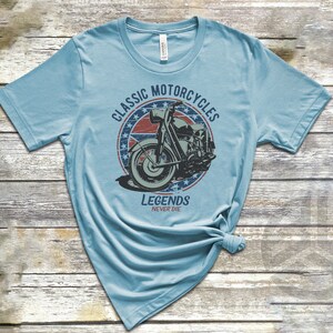 Op de afbeelding: Een lichtblauw t-shirt met een vintage-stijl afbeelding van een motorfiets. De afbeelding heeft een rood, wit en blauw sterrendesign en de tekst "Classic Motorcycles Legends Never Die".