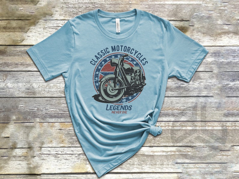 Op de afbeelding: Een lichtblauw t-shirt met een vintage-stijl afbeelding van een motorfiets. De afbeelding heeft een rood, wit en blauw sterrendesign en de tekst "Classic Motorcycles Legends Never Die".