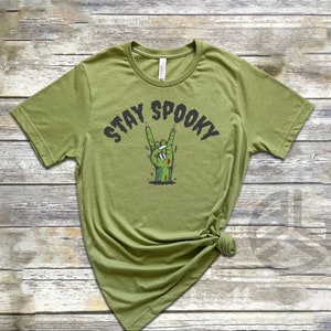 Puede incluir: Una camiseta verde con el texto "Stay Spooky" en letras negras. Una mano de zombie verde con u&ntilde;as negras est&aacute; haciendo el gesto de rock and roll.