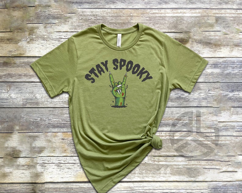 Puede incluir: Una camiseta verde con el texto "Stay Spooky" en letras negras. Una mano de zombie verde con u&ntilde;as negras est&aacute; haciendo el gesto de rock and roll.