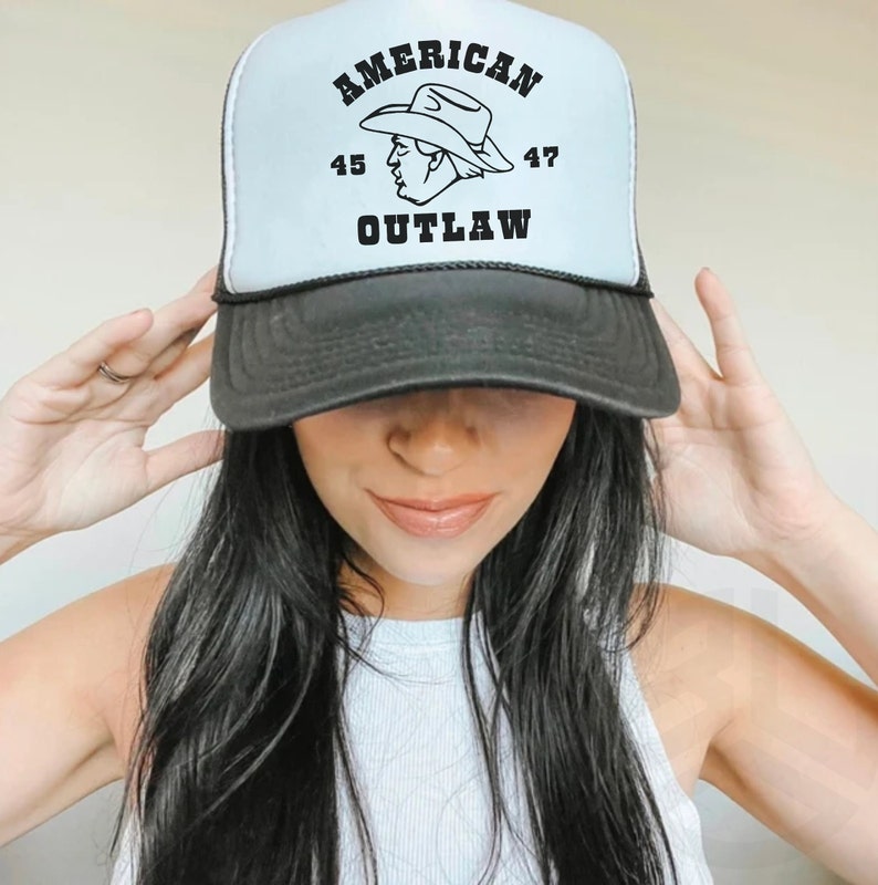 May include: A white trucker hat with a black mesh back. The front of the hat has black text that reads "AMERICAN 45 47 OUTLAW" and a black and white illustration of a person wearing a cowboy hat.
