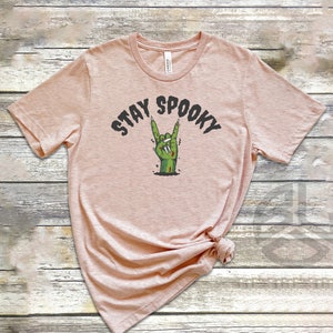 Puede incluir: Una camiseta rosa claro con el texto "STAY SPOOKY" en letras negras y una mano de zombie verde haciendo un gesto de rock and roll.