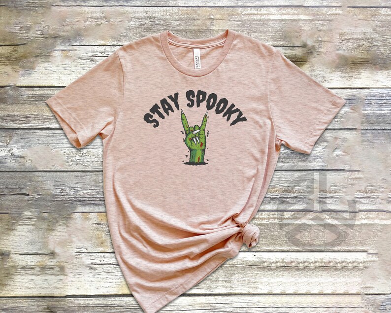 Puede incluir: Una camiseta rosa claro con el texto "STAY SPOOKY" en letras negras y una mano de zombie verde haciendo un gesto de rock and roll.
