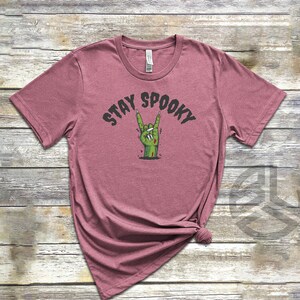 Puede incluir: Una camiseta de color malva con un gr&aacute;fico de una mano de zombi haciendo el gesto de rock and roll. El texto "Stay Spooky" est&aacute; impreso sobre la mano.