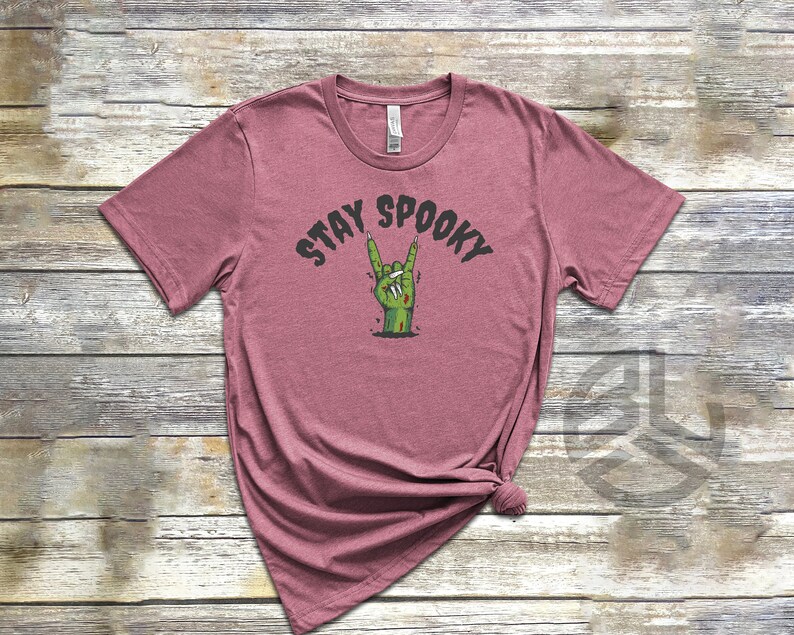 Puede incluir: Una camiseta de color malva con un gr&aacute;fico de una mano de zombi haciendo el gesto de rock and roll. El texto "Stay Spooky" est&aacute; impreso sobre la mano.