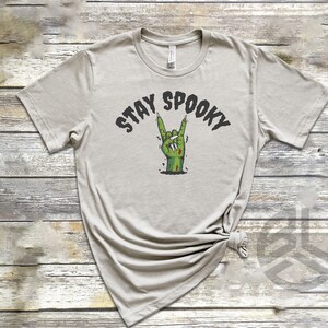 Puede incluir: Una camiseta gris claro con el texto "Stay Spooky" en letras negras. Una mano de zombie verde con u&ntilde;as rojas est&aacute; haciendo el gesto de rock and roll.