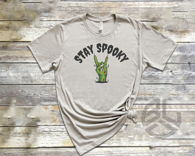 Puede incluir: Una camiseta gris claro con el texto "Stay Spooky" en letras negras. Una mano de zombie verde con u&ntilde;as rojas est&aacute; haciendo el gesto de rock and roll.