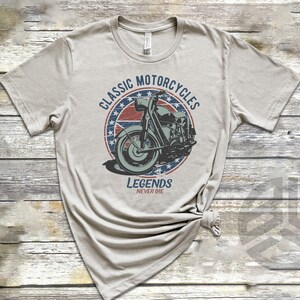 Op de afbeelding: Een lichtgrijs T-shirt met een vintage-stijl grafische afbeelding van een motorfiets in een cirkel met rode, witte en blauwe sterren. De tekst "Classic Motorcycles" staat boven de motorfiets en "Legends Never Die" staat onder de motorfiets.