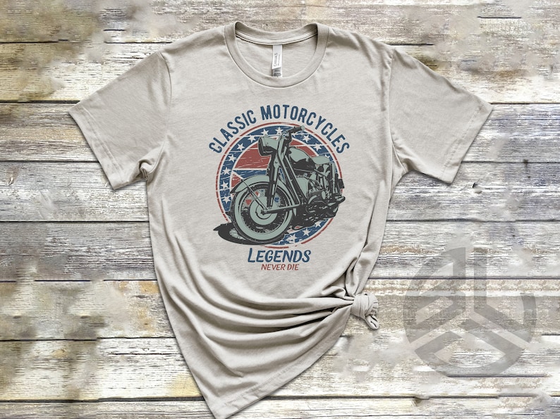 Op de afbeelding: Een lichtgrijs T-shirt met een vintage-stijl grafische afbeelding van een motorfiets in een cirkel met rode, witte en blauwe sterren. De tekst "Classic Motorcycles" staat boven de motorfiets en "Legends Never Die" staat onder de motorfiets.