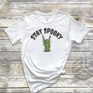 Puede incluir: Camiseta blanca con una mano de zombie verde haciendo el signo de rock and roll. El texto "Stay Spooky" est&aacute; por encima de la mano.